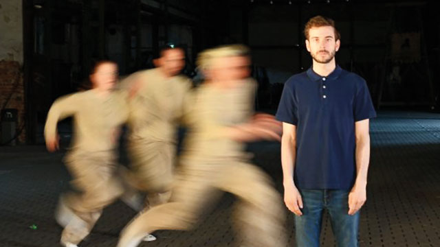 ein junger Mann steht auf einem Platz, hinter ihm laufen drei nicht erkennbare, verschwommene Menschen