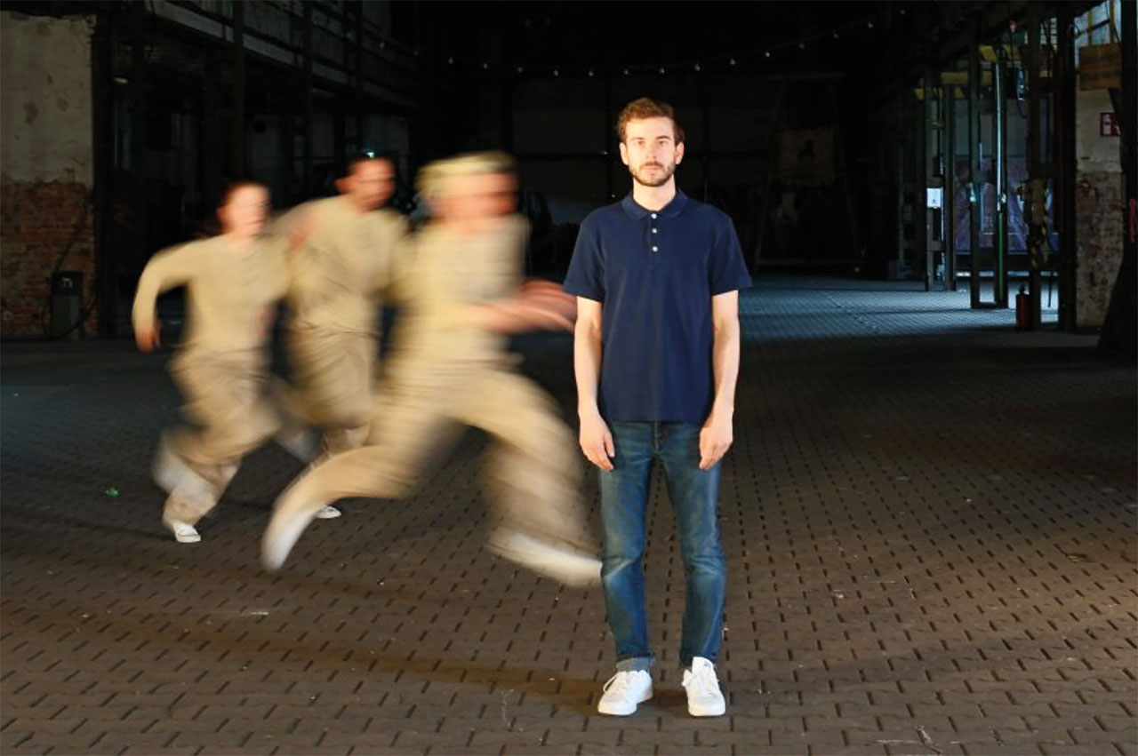 ein junger Mann steht auf einem Platz, hinter ihm laufen drei nicht erkennbare, verschwommene Menschen